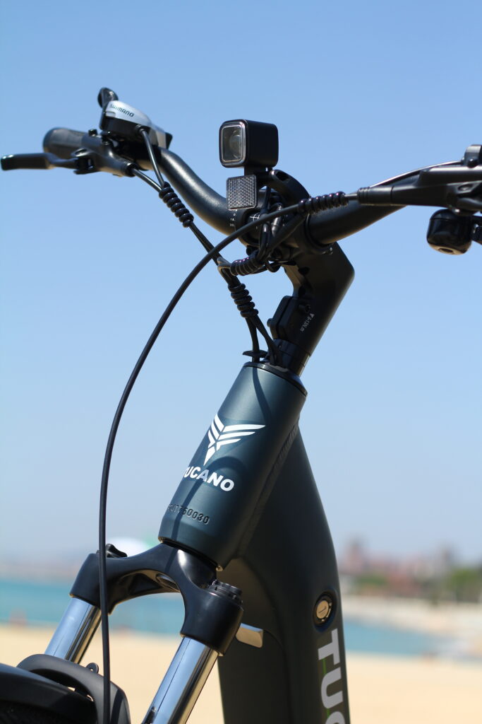 Mujer sonriente en bicicleta eléctrica Tucano, en un paisaje urbano con edificios modernos. Refleja un estilo de vida activo y sostenible en la ciudad.
