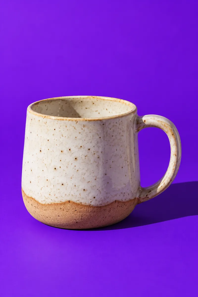 Taza de cerámica artesanal esmaltada en blanco moteado sobre un fondo liso morado con sombra dura