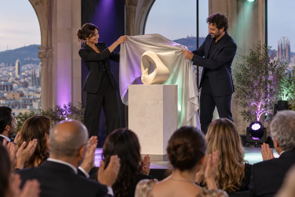 Dos presentadores retiran la tela que cubre un objeto de diseño sobre una peana mientras el público aplaude bajo las luces del escenario